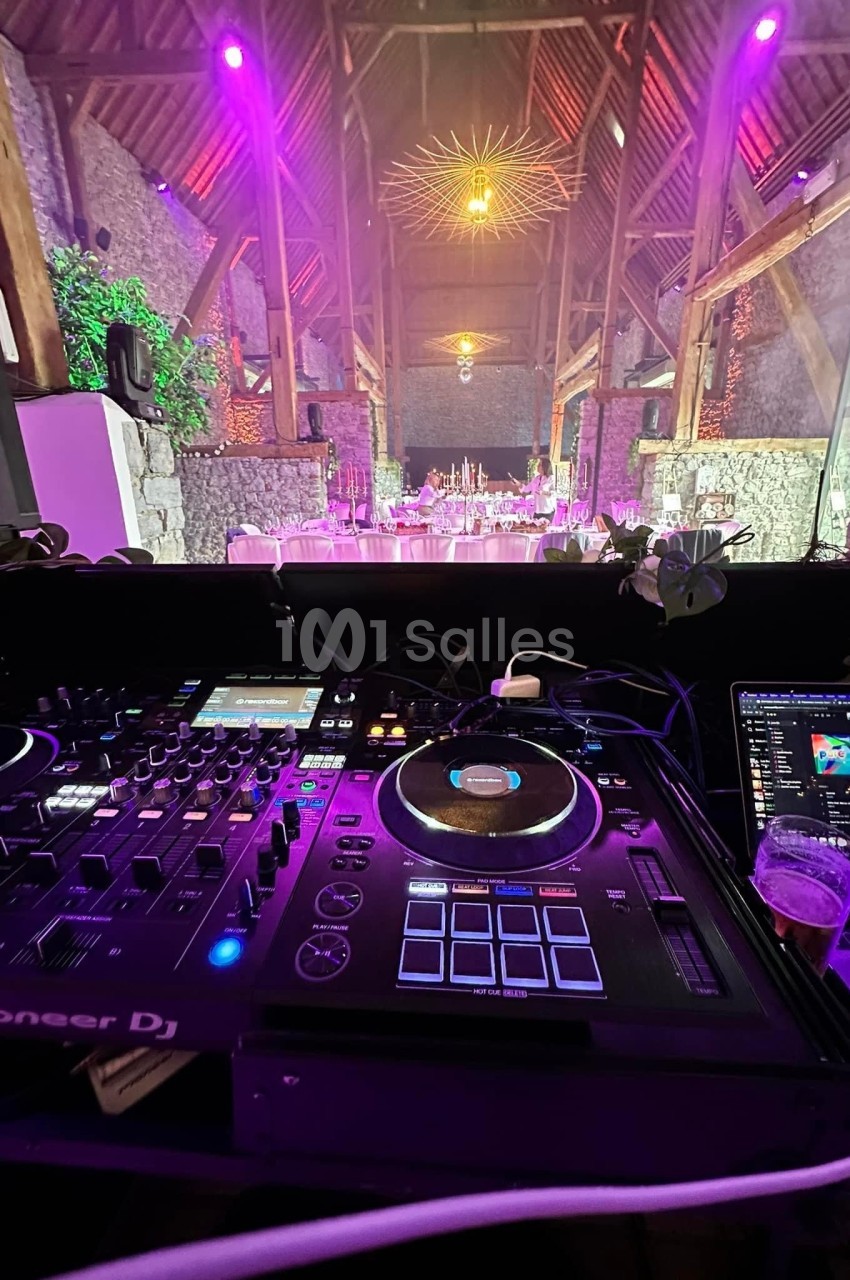 Vue depuis une table de mixage DJ dans une salle en pierre éclairée de lumières violettes, avec des tables dressées au fond.