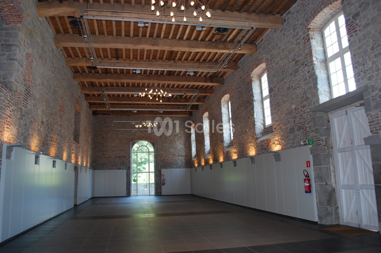Grande salle en pierre avec plafond en bois, fenêtres cintrées, éclairage suspendu et porte vitrée donnant sur l'extérieur.