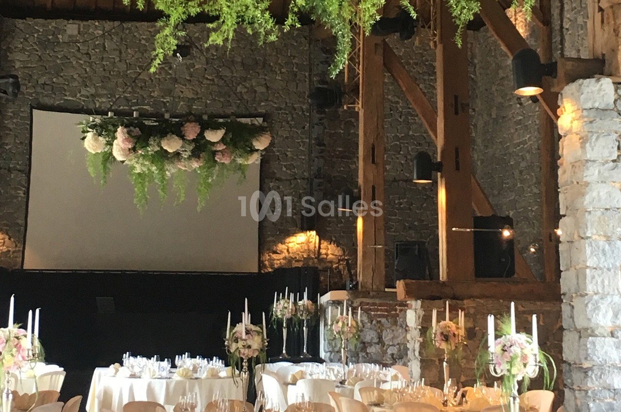 Salle de réception en pierre avec tables décorées de fleurs et chandeliers, et une scène ornée de verdure suspendue.
