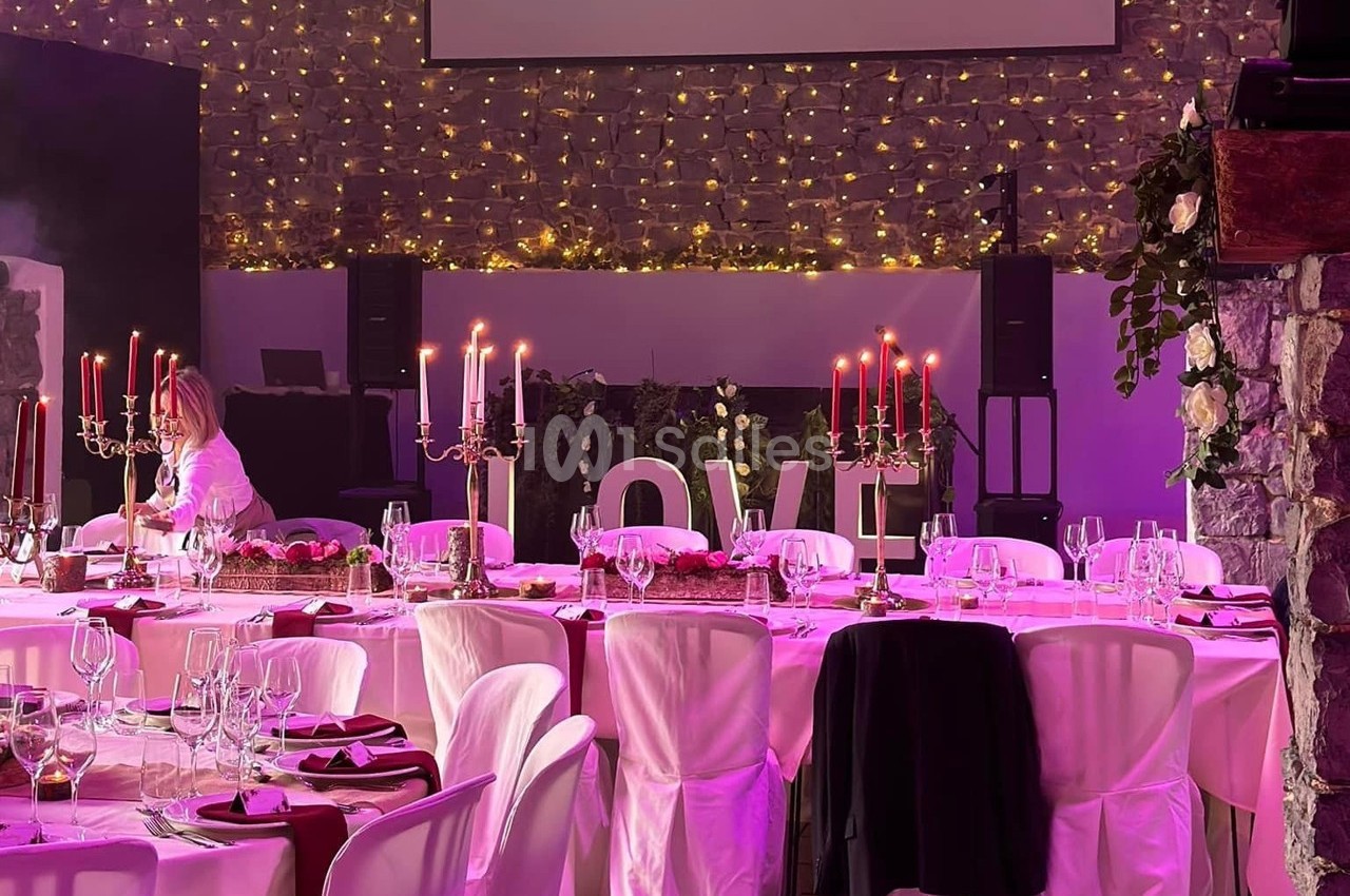 Salle de réception décorée pour un événement, avec tables dressées, chandeliers et guirlandes lumineuses en arrière-plan.