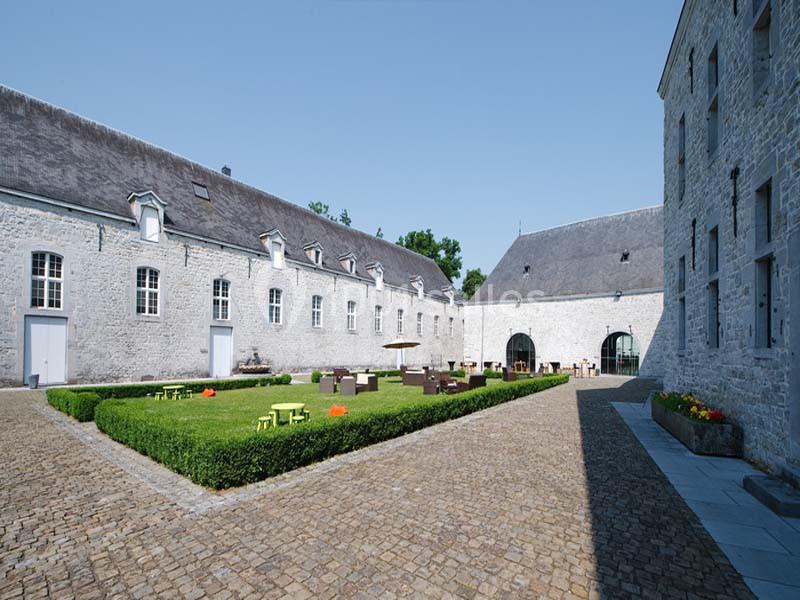 Cour intérieure pavée entourée de bâtiments en pierre, avec des tables et des chaises disposées près d'un jardin central.