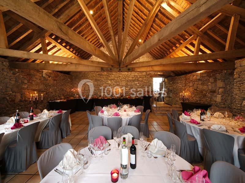 Salle en pierre avec poutres apparentes, tables rondes dressées avec nappes grises et serviettes roses, ambiance chaleureuse.