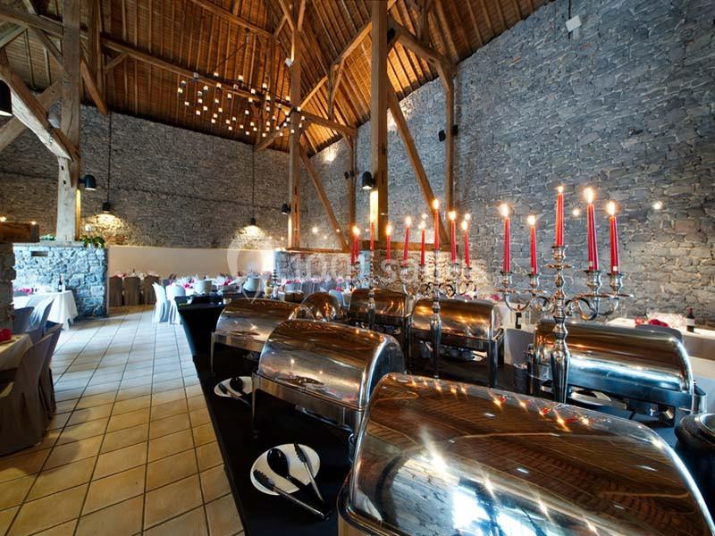 Salle de réception en pierre avec plafond en bois, tables dressées et buffets en inox ornés de chandeliers rouges.