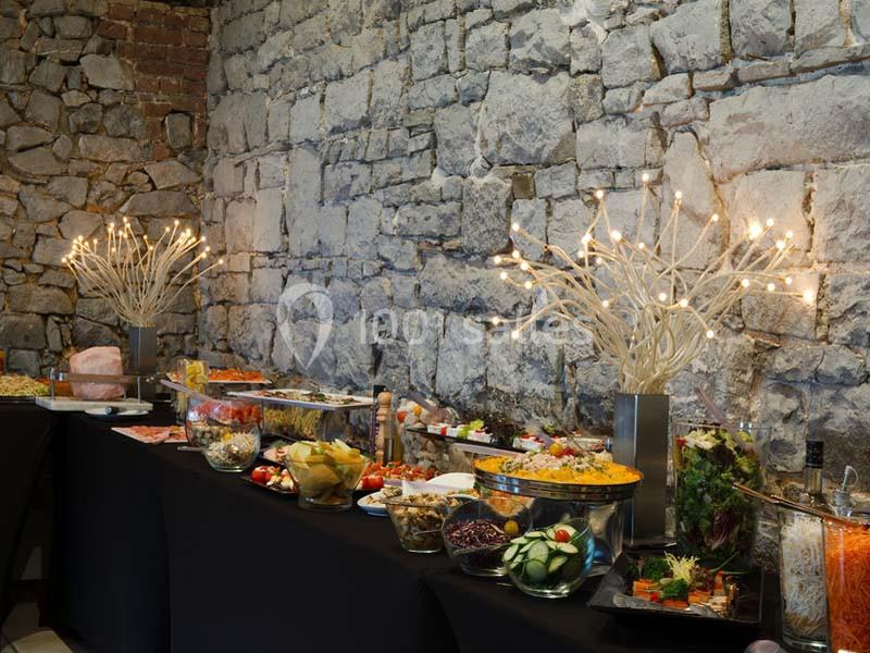 Buffet varié disposé sur des tables noires, avec un mur en pierre en arrière-plan et des décorations lumineuses.