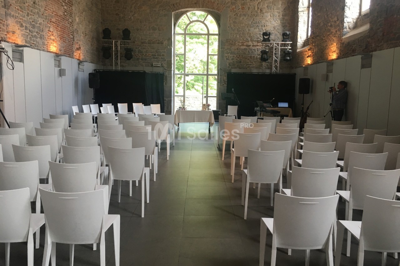 Salle en pierre avec des rangées de chaises blanches alignées face à une table et une grande fenêtre en arrière-plan.