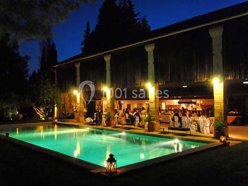 Vue nocturne d'une piscine éclairée devant une salle de réception animée, entourée de végétation.