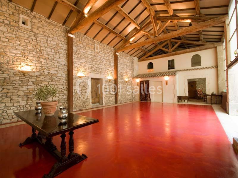 Salle spacieuse avec murs en pierre apparente, plafond en poutres de bois et sol rouge brillant, éclairée par des appliques…