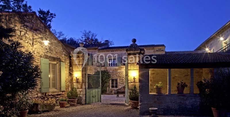 Cour intérieure d'une maison en pierre éclairée par des lampes extérieures, avec un portail ouvert au crépuscule.
