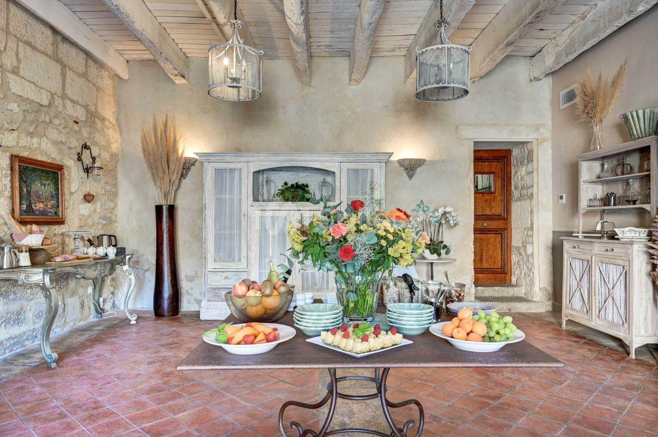 Table dressée avec fruits frais et fleurs dans une cuisine rustique lumineuse aux murs en pierre et poutres apparentes.