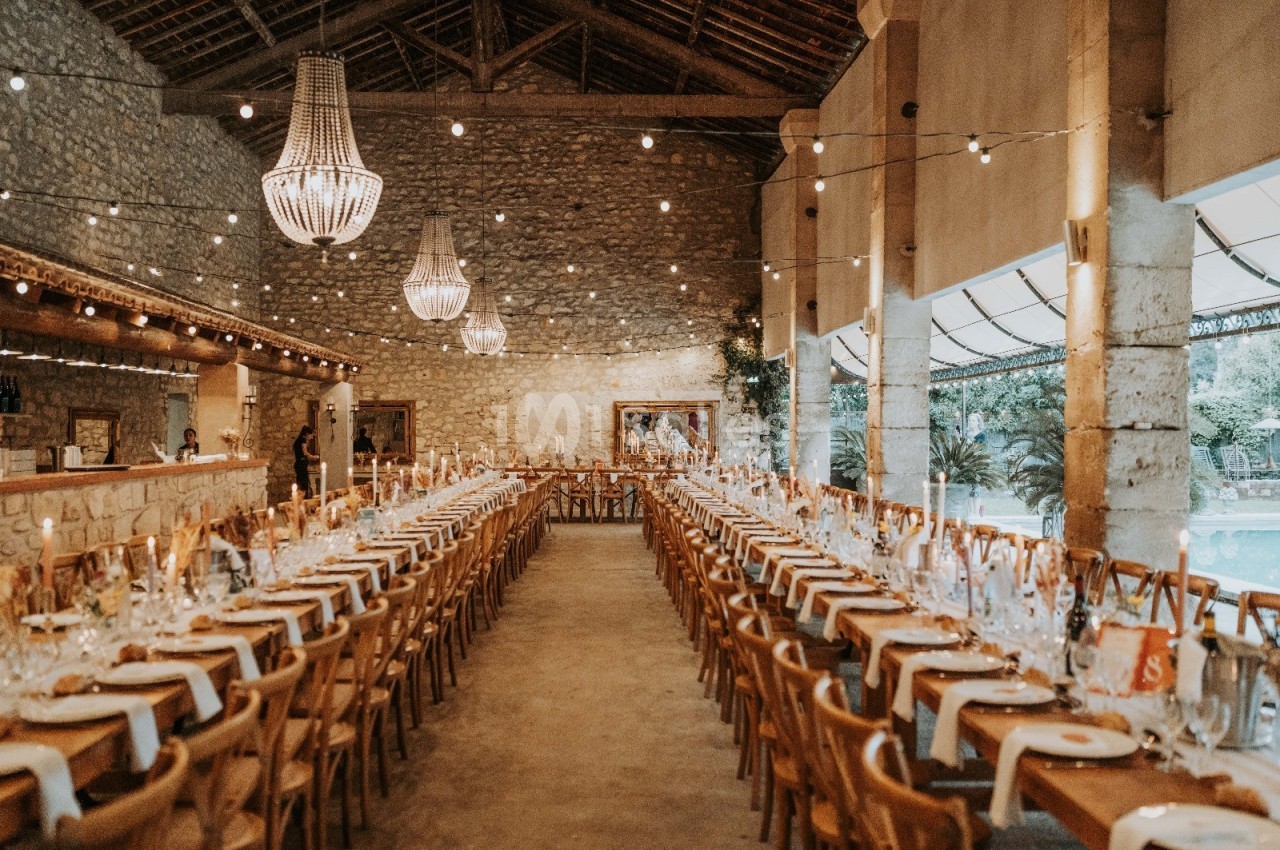 Grande salle en pierre avec de longues tables dressées pour un banquet, éclairée par des lustres et des guirlandes…