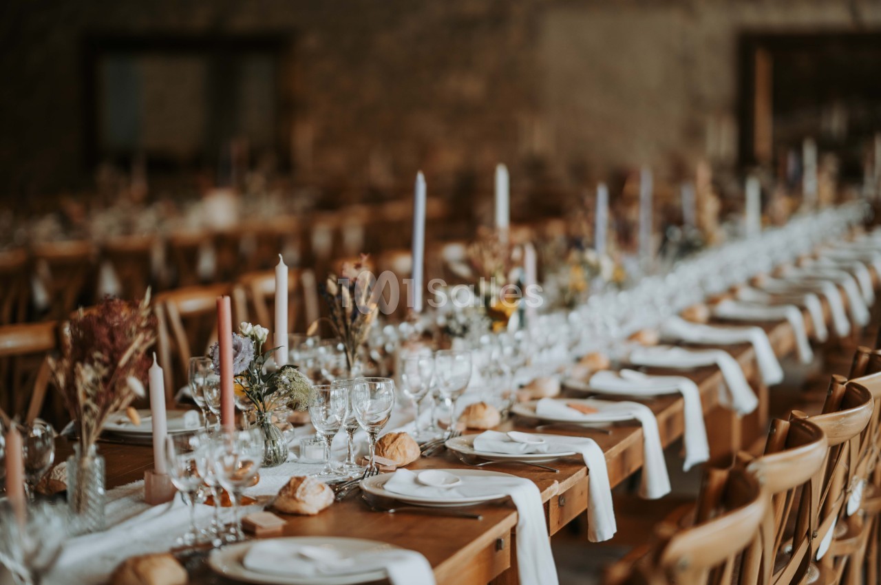 Grande table de banquet décorée avec des fleurs séchées, des bougies et des couverts alignés dans une salle rustique.