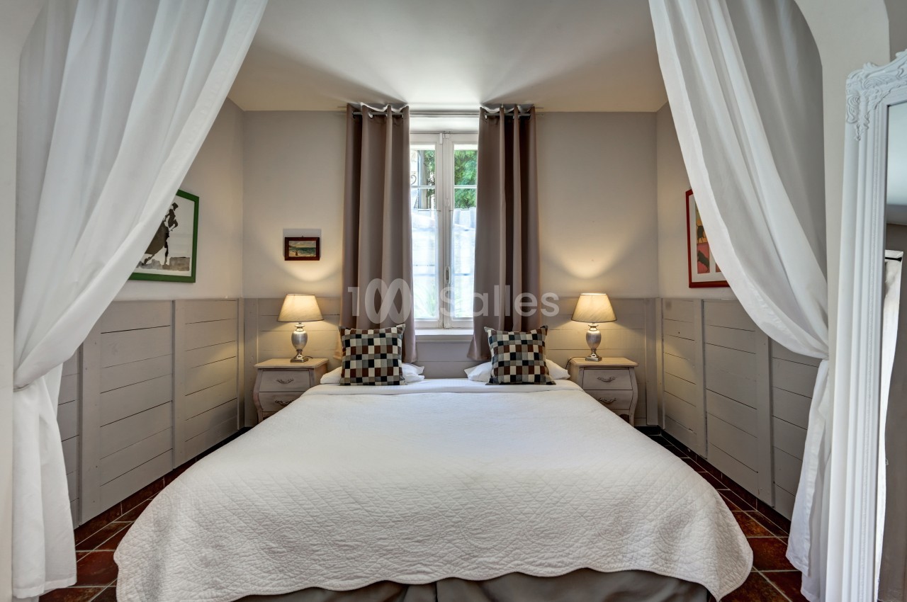 Lit double avec couvre-lit blanc, encadré de rideaux ouverts, deux lampes de chevet allumées et une fenêtre en arrière-plan.