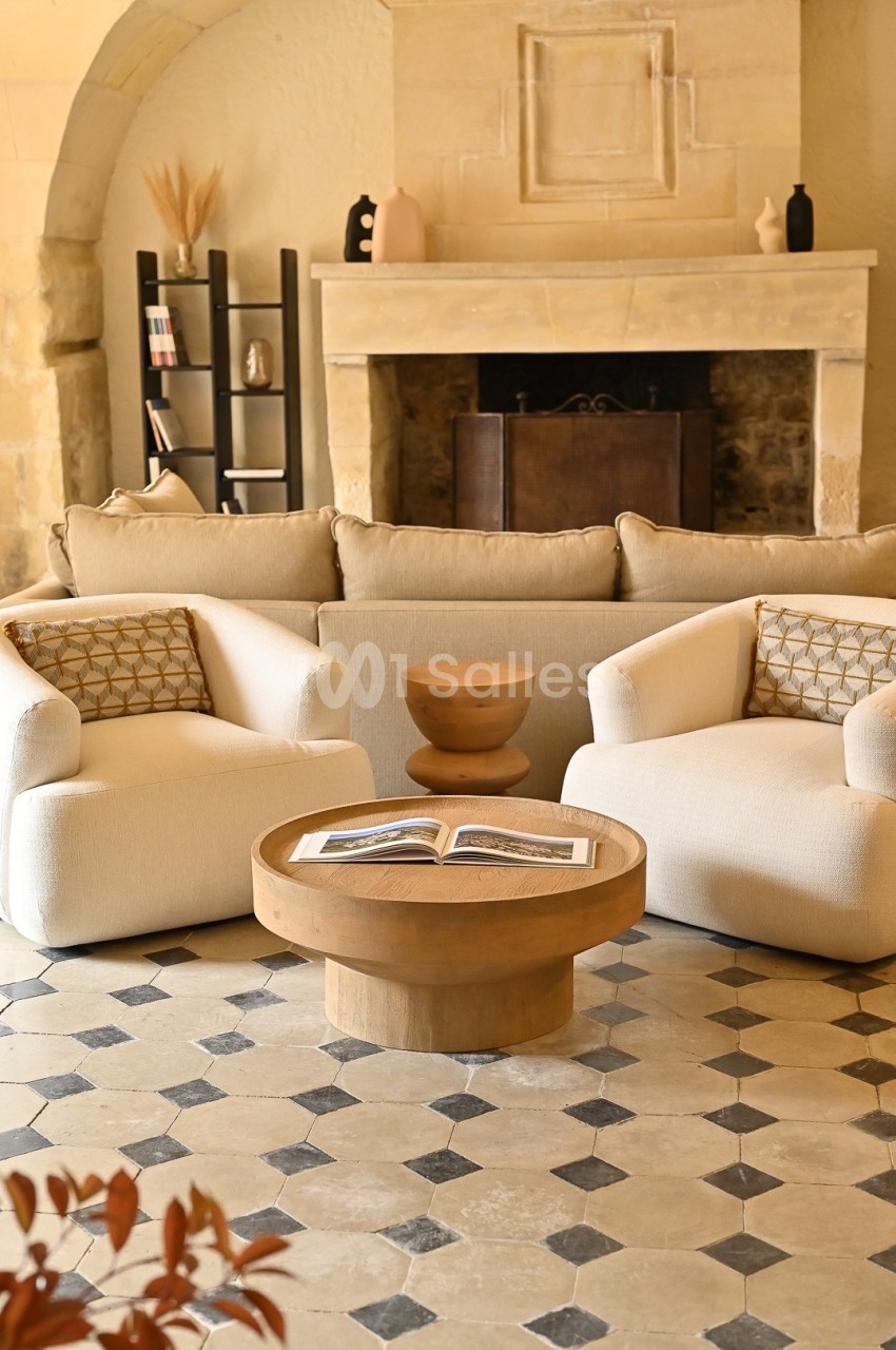 Salon chaleureux avec cheminée en pierre, canapé beige, fauteuils blancs, table basse en bois et sol en carreaux.