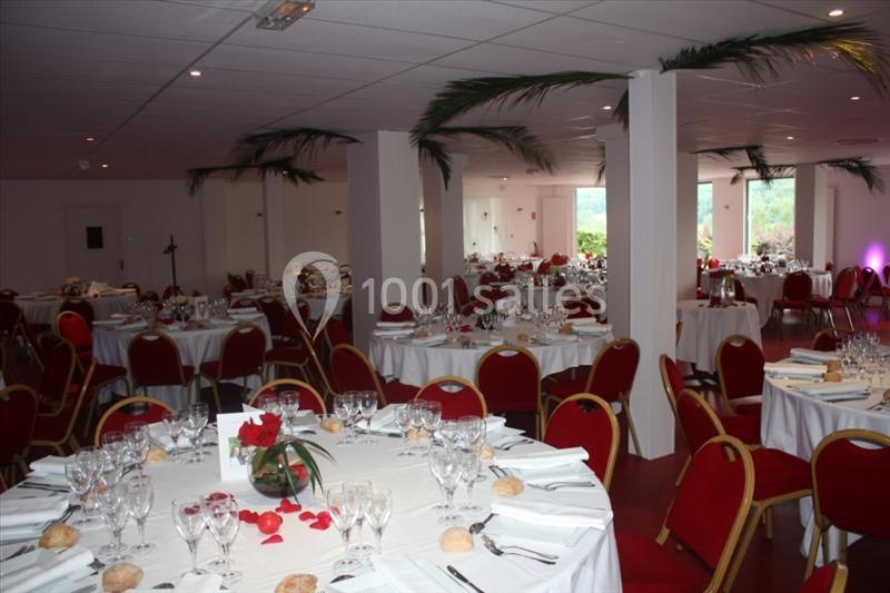 Salle de réception avec tables rondes dressées, nappes blanches, chaises rouges et décorations florales.