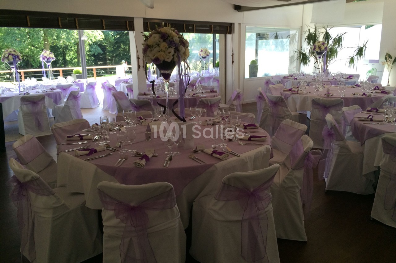 Salle de réception décorée pour un événement, avec des tables rondes, nappes blanches et ornements violets.