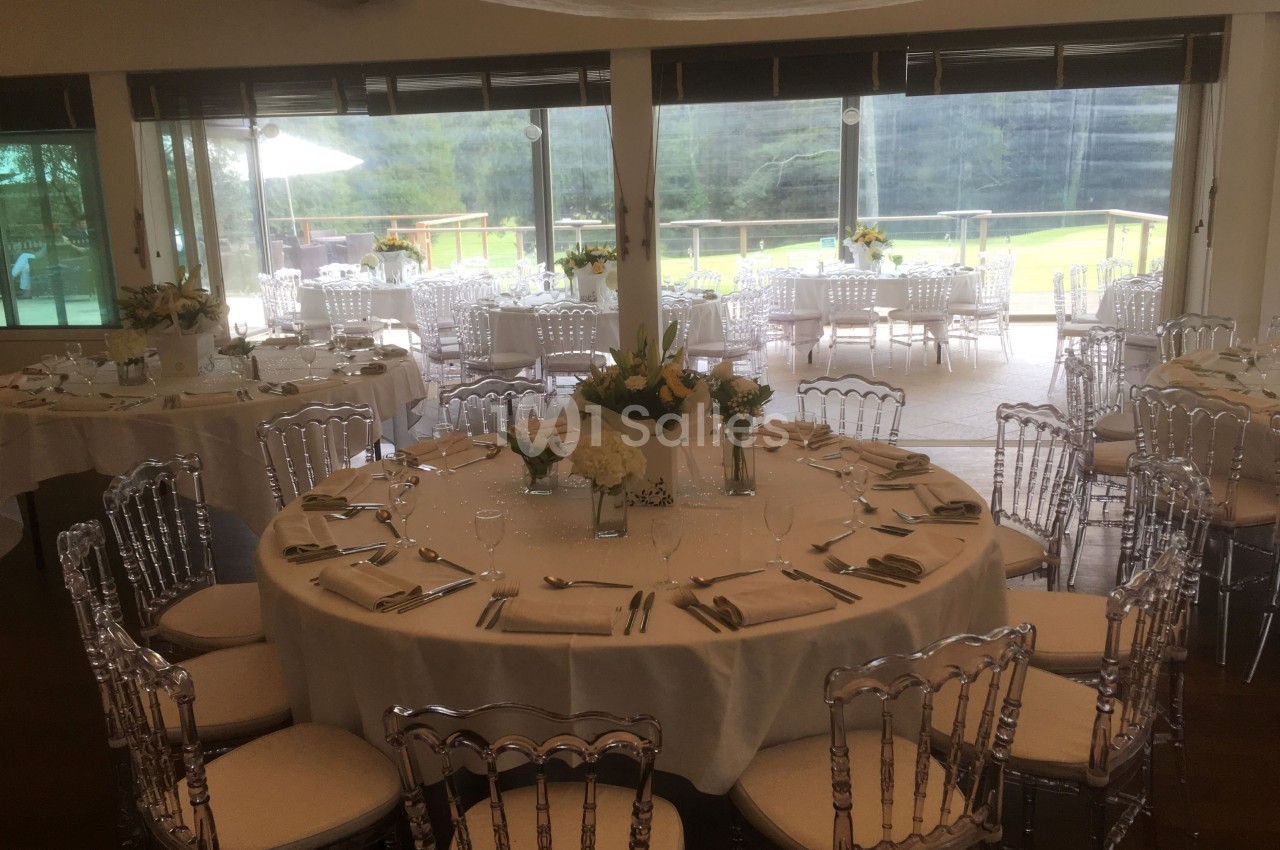 Salle de réception lumineuse avec tables rondes dressées, chaises transparentes et vue sur un espace extérieur verdoyant.