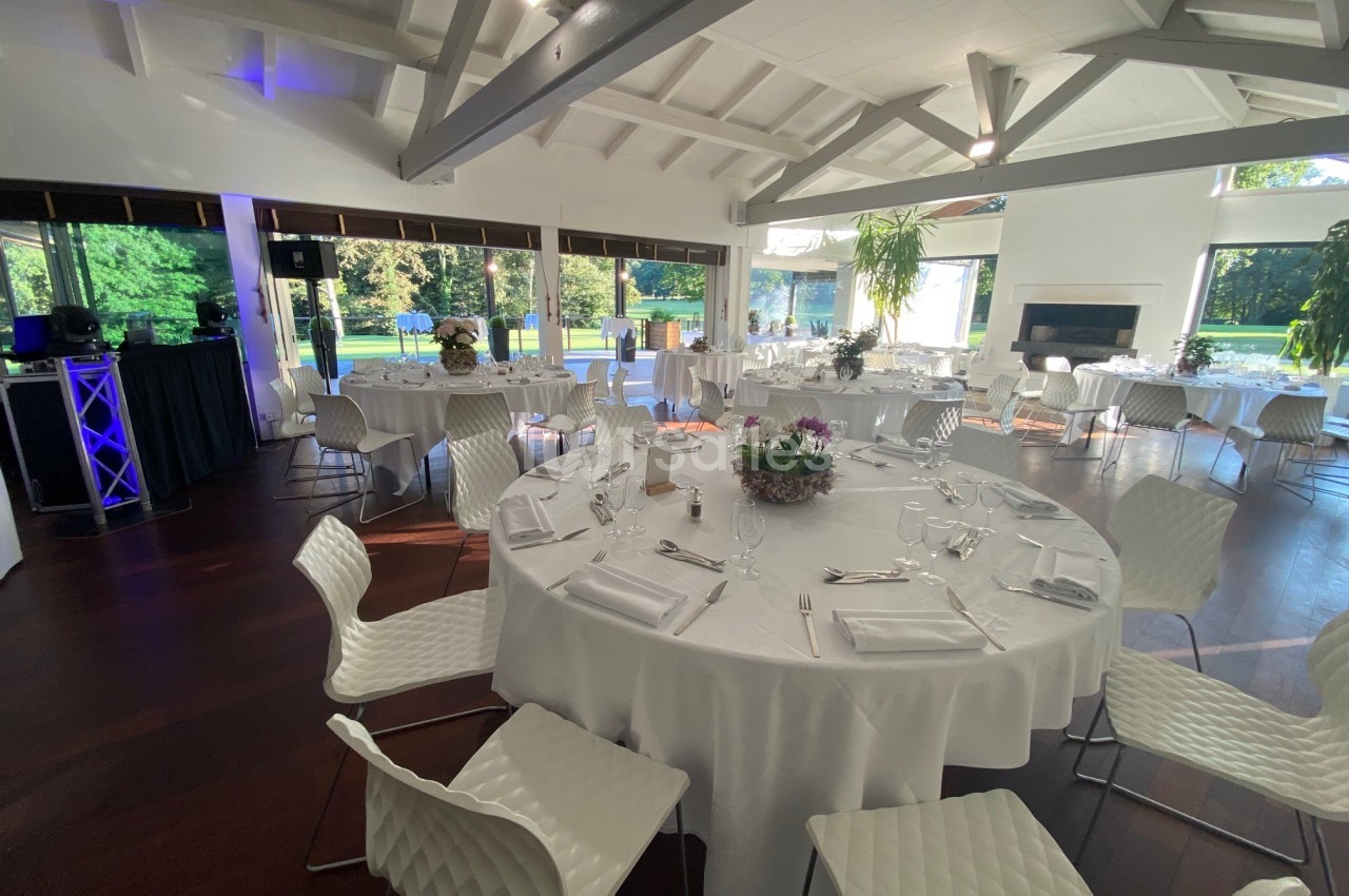 Salle de réception lumineuse avec tables rondes dressées, chaises blanches et vue sur un jardin verdoyant.