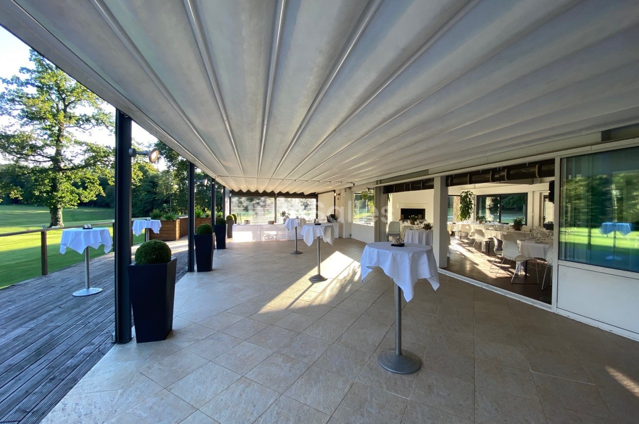 Terrasse couverte avec tables hautes dressées, vue sur un jardin verdoyant et grandes baies vitrées.