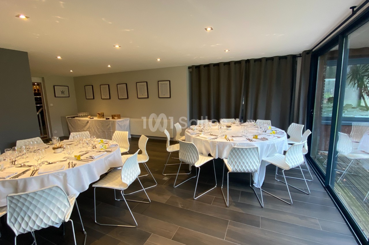 Salle lumineuse avec tables rondes dressées pour un repas, chaises blanches et grandes baies vitrées donnant sur l'extérieur.