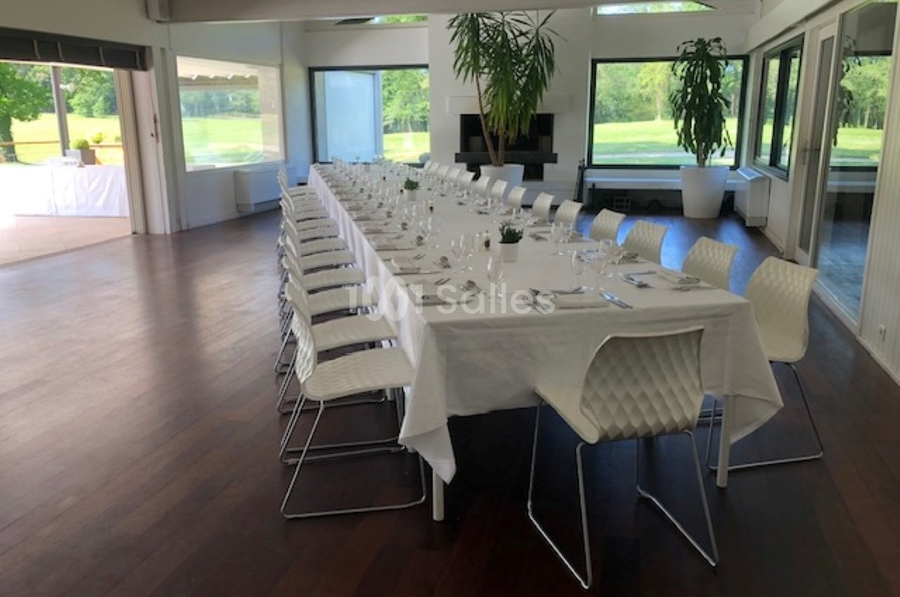Salle lumineuse avec une grande table rectangulaire dressée pour un repas, entourée de chaises blanches modernes.