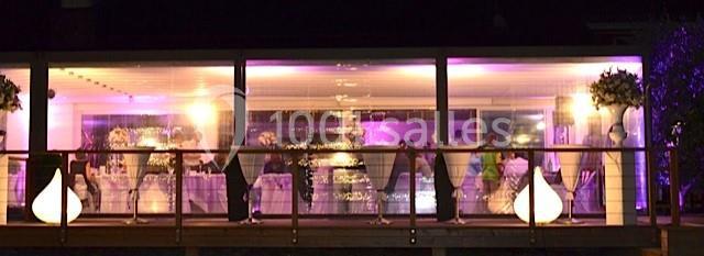 Salle vitrée éclairée en violet, avec des invités debout près de tables décorées, vue de l'extérieur.