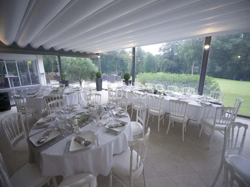 Salle de réception lumineuse avec tables rondes dressées, chaises blanches et vue sur un jardin verdoyant.