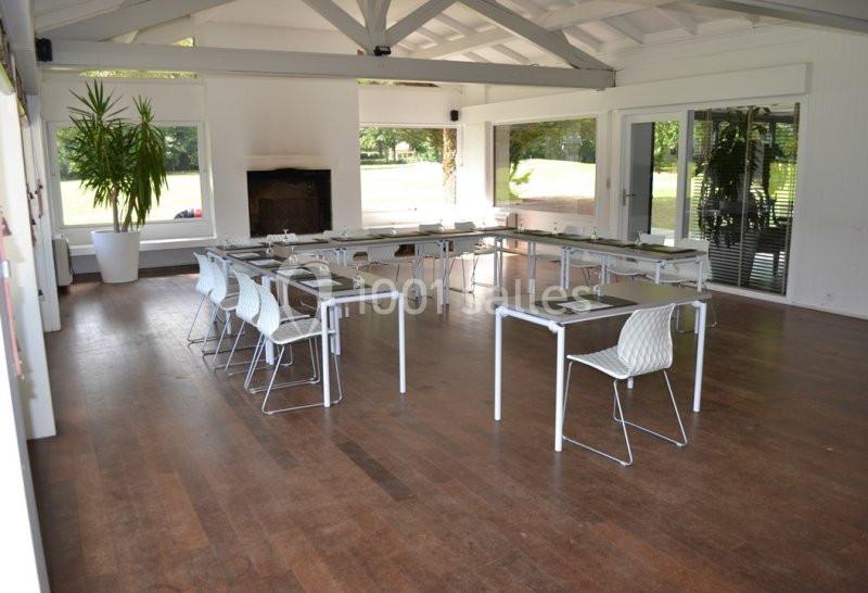 Salle lumineuse aménagée en U avec des tables blanches, des chaises et une vue sur un espace extérieur verdoyant.