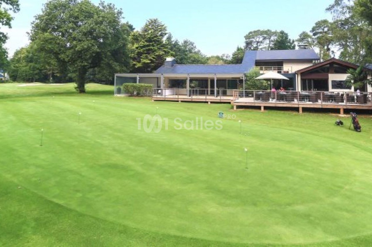 Terrain de golf avec pelouse bien entretenue, entouré d'arbres, et un club-house avec terrasse en arrière-plan.
