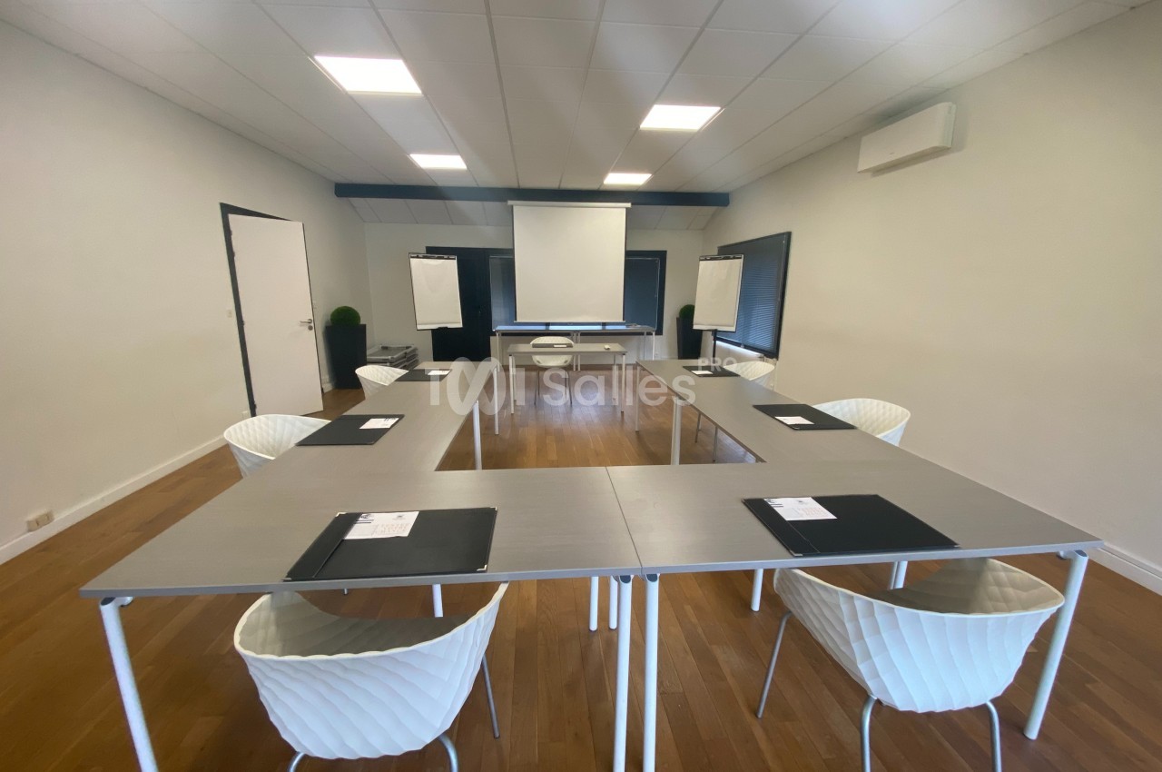 Salle de réunion lumineuse avec tables disposées en U, chaises blanches, écran de projection et tableaux blancs.