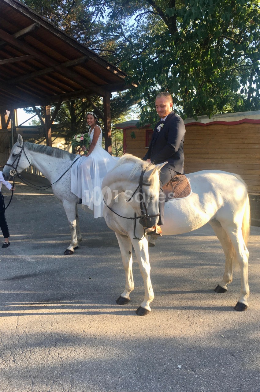 Un couple en tenue de mariage est assis sur deux chevaux blancs, accompagné d'une personne tenant un cheval.