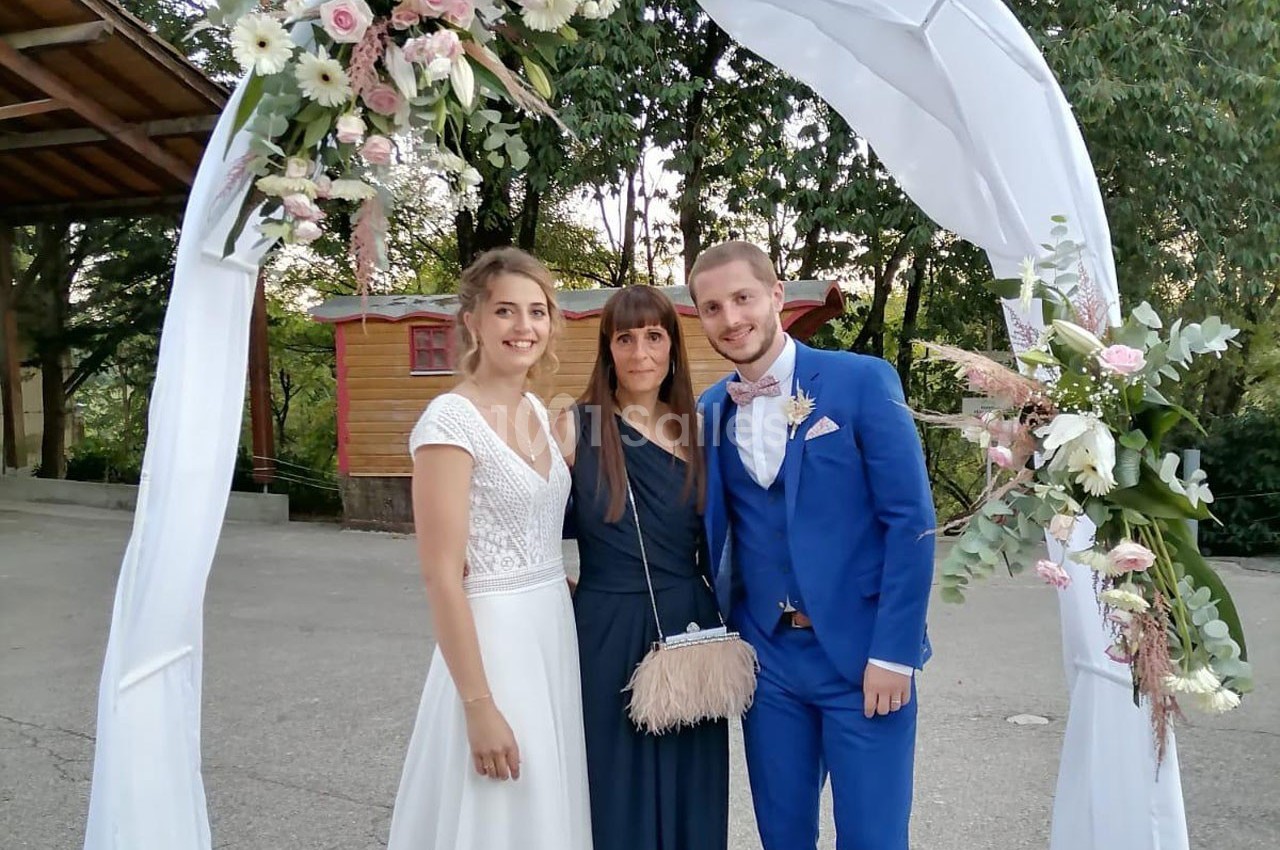 Un couple en tenue de mariage pose avec une femme sous une arche décorée de fleurs, en extérieur.