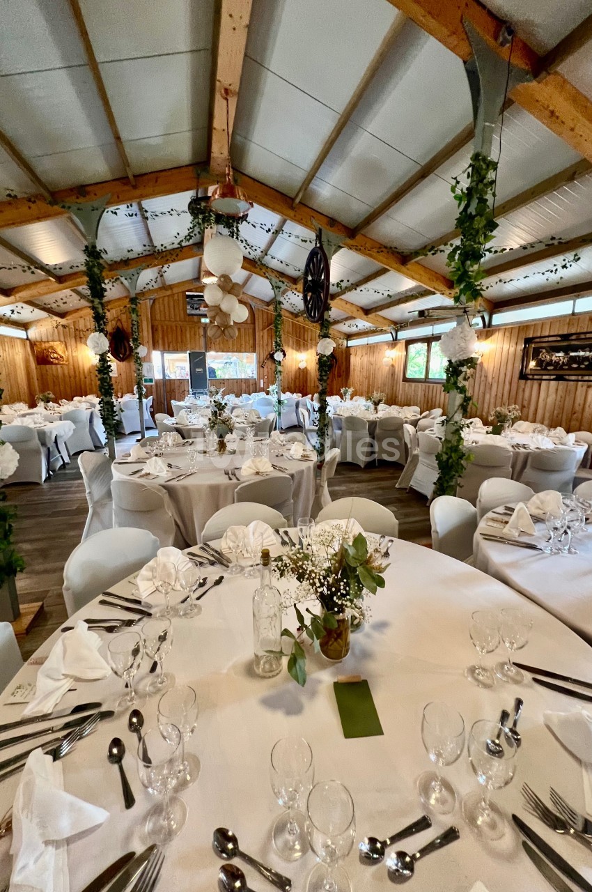 Salle de réception décorée avec des tables rondes dressées, ornées de fleurs et entourées de chaises blanches.