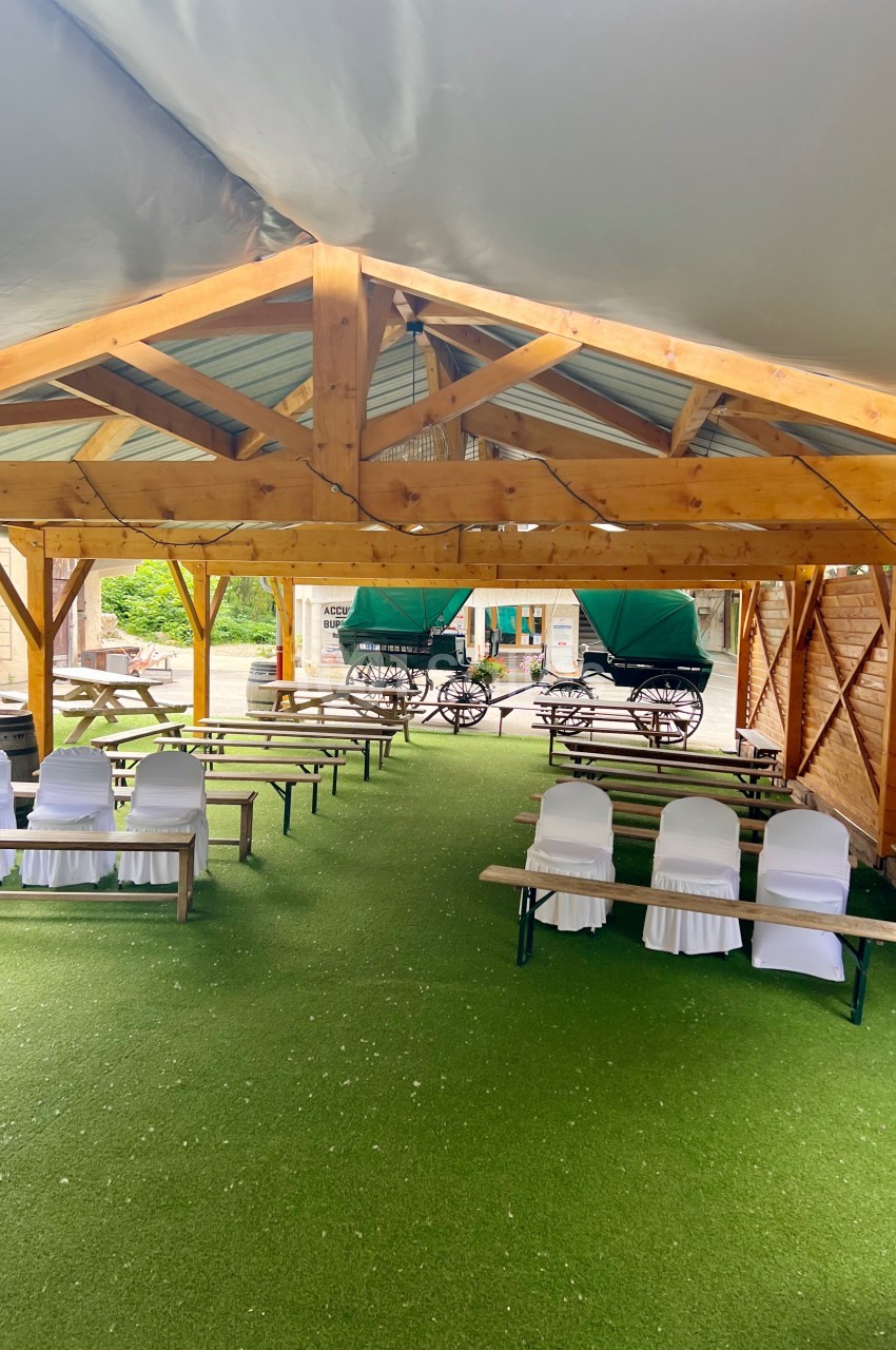 Structure en bois abritant des bancs et des chaises blanches sur un sol en gazon synthétique, avec des calèches en arrière…