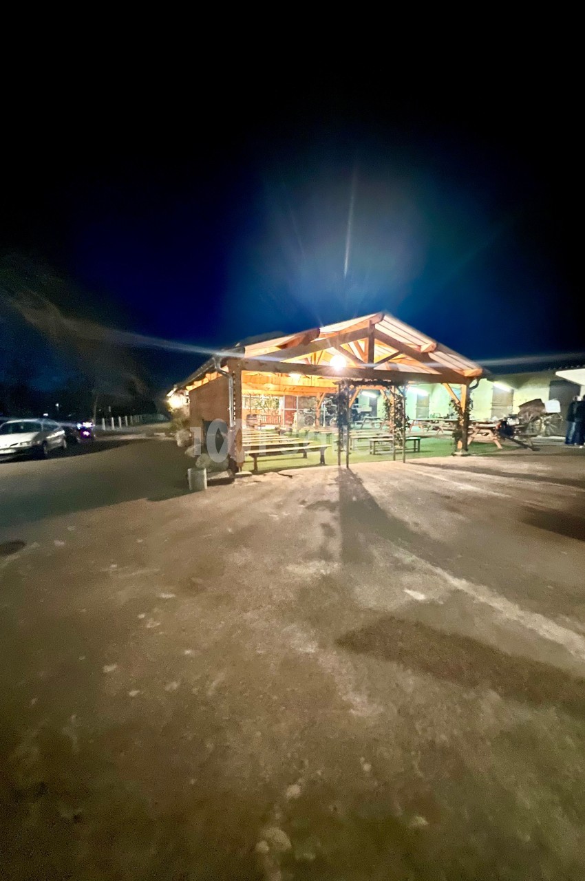 Abri en bois éclairé la nuit, entouré de bancs et situé sur un parking avec quelques voitures visibles.