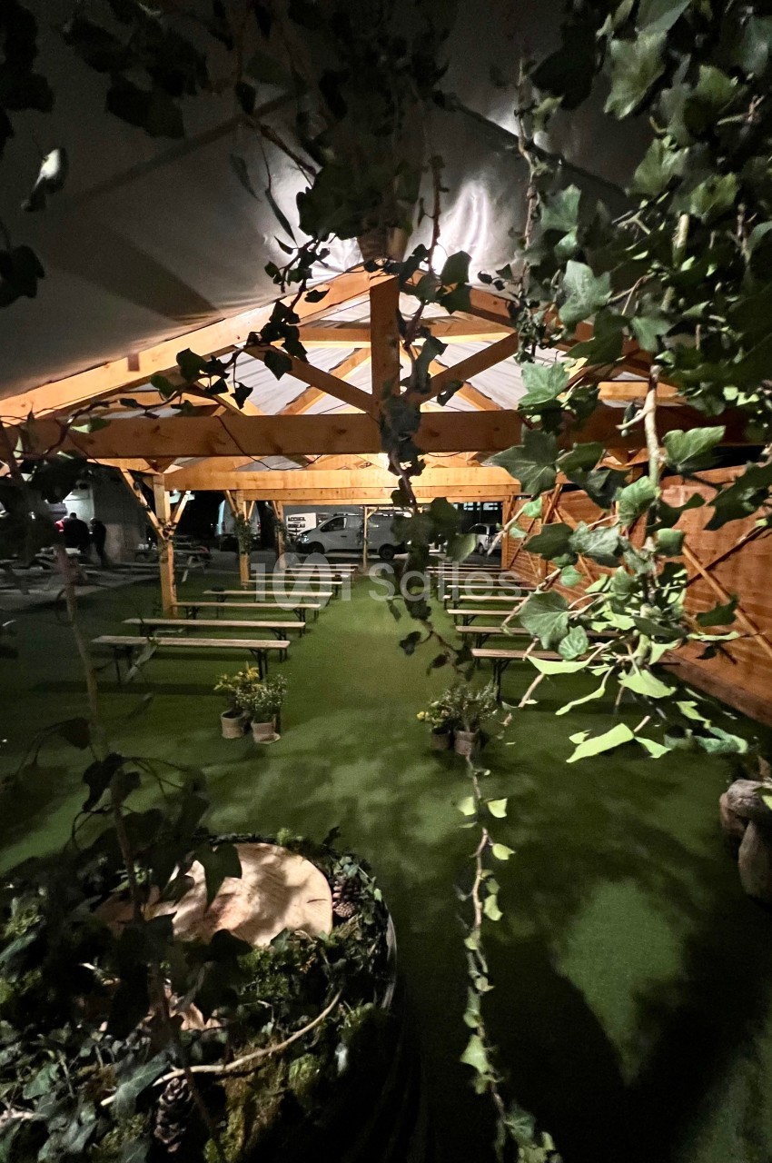 Structure en bois avec bancs alignés sous une tente, entourée de végétation et éclairée par une lumière douce.