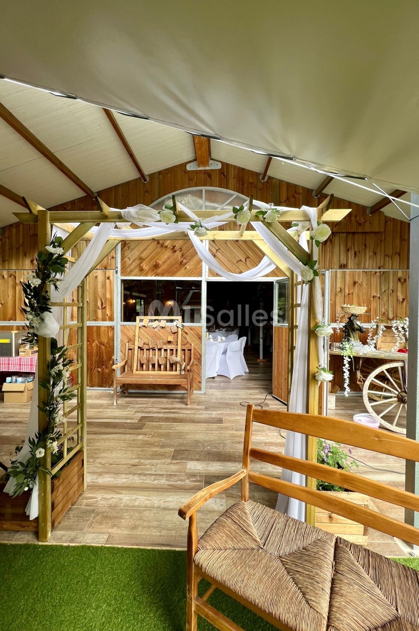 Arche décorée de fleurs et de tissu blanc devant une salle en bois aménagée pour un événement festif.