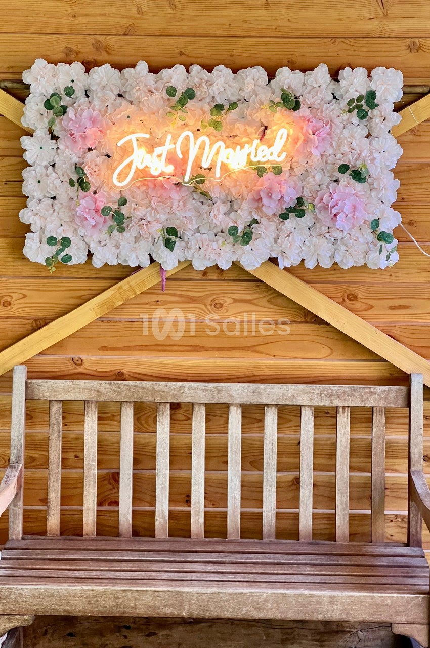 Banc en bois devant un panneau floral avec une inscription lumineuse ’Just Married’ sur un mur en bois.