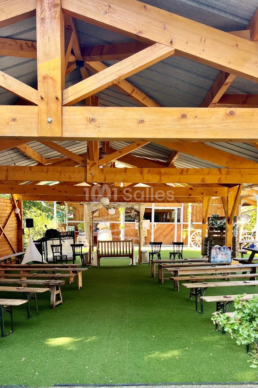 Structure en bois avec toit en métal abritant des bancs et des décorations, entourée de verdure et de fleurs.