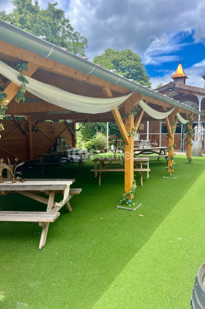 Abri en bois décoré de guirlandes et nappes blanches, avec tables de pique-nique sur une pelouse synthétique.