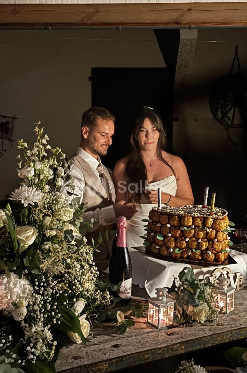 Deux mariés devant une table décorée de fleurs blanches, gâteau de choux et bouteille de champagne.
