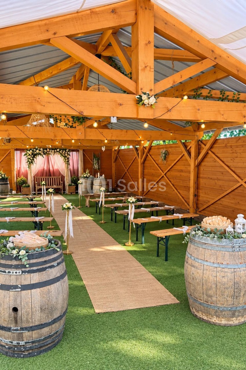 Chapiteau en bois avec charpente apparente, tables pliantes dressées sur gazon artificiel, tonneaux fleuris, guirlande lumineuse et fond de scène en tissu rose.