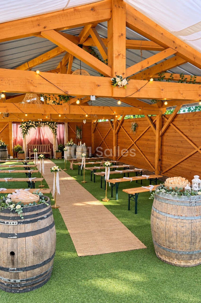 Structure en bois avec poutres apparentes, sol en gazon synthétique, tables pliantes dressées avec décoration florale, tonneaux de bois aux extrémités et allée centrale.