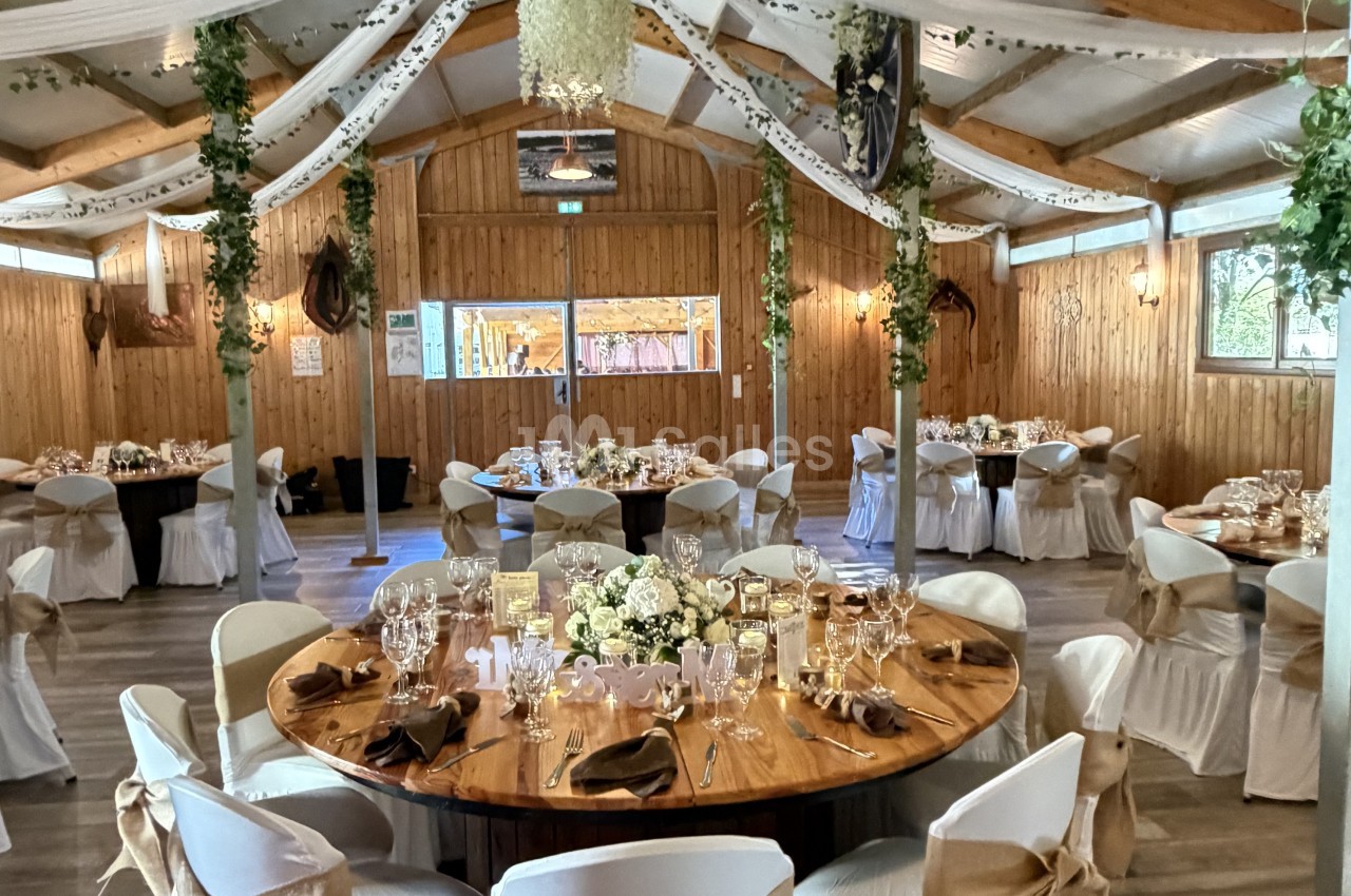 Salle rustique en bois avec tables rondes dressées, chaises blanches, décoration florale blanche et guirlandes vertes au plafond.