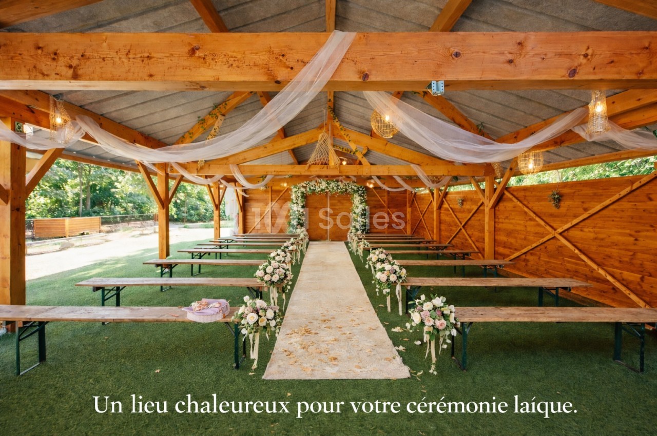 Grange en bois avec poutres apparentes, allée centrale drapée de tissu blanc, bancs décorés de fleurs blanches et roses.