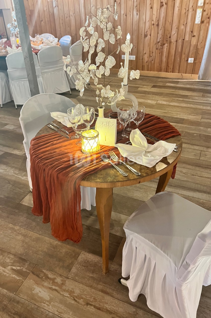 Table ronde décorée pour un dîner, avec nappe orange, vaisselle élégante, bougie allumée et centre de table floral.