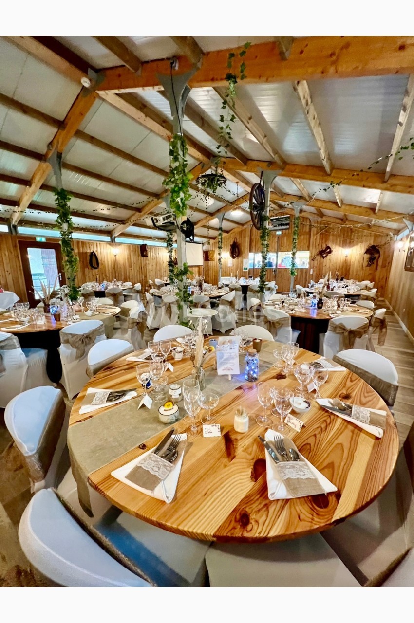 Salle de réception décorée avec des tables rondes en bois, nappes blanches et éléments végétaux suspendus.