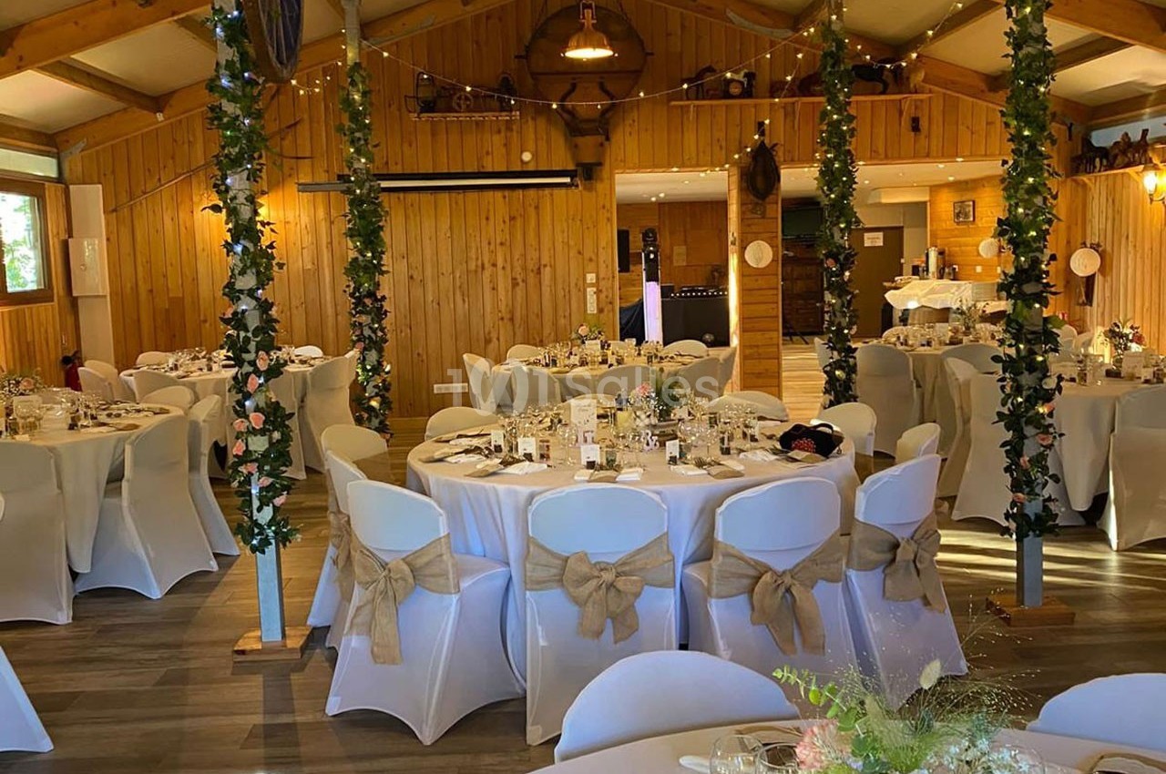 Salle de réception décorée avec des tables rondes, nappes blanches, chaises ornées de rubans et colonnes fleuries.