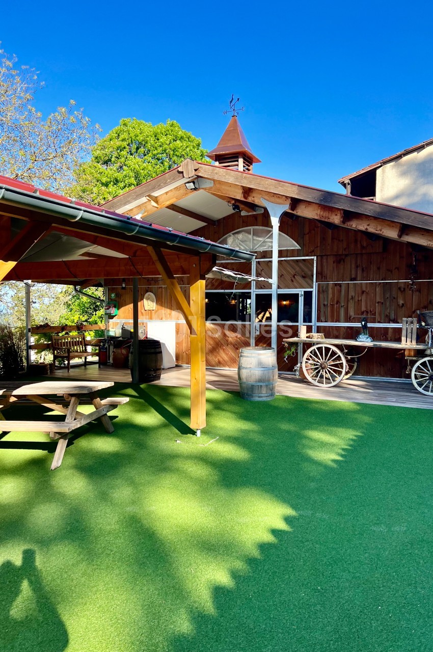 Cour extérieure avec gazon synthétique, tables en bois, charrette décorative et bâtiment en bois sous un ciel bleu.