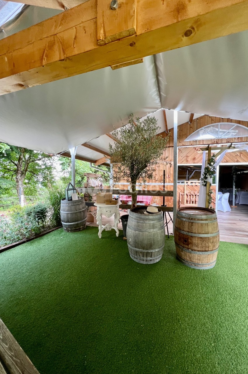 Terrasse couverte avec sol en gazon synthétique, tonneaux en bois, table blanche et arbre décoratif.