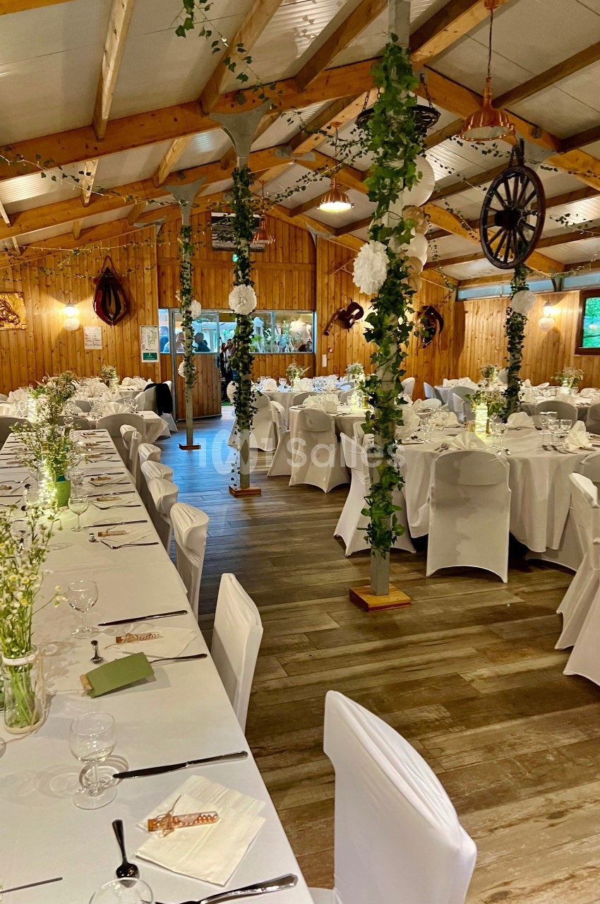 Salle de réception en bois décorée avec des tables dressées, des chaises blanches et des guirlandes végétales.